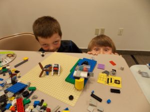 Two children peek from behind a table at Lego Club, summer 2016