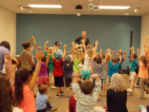 Children and family members raise their hands in the air during a concert with Andy Mason.