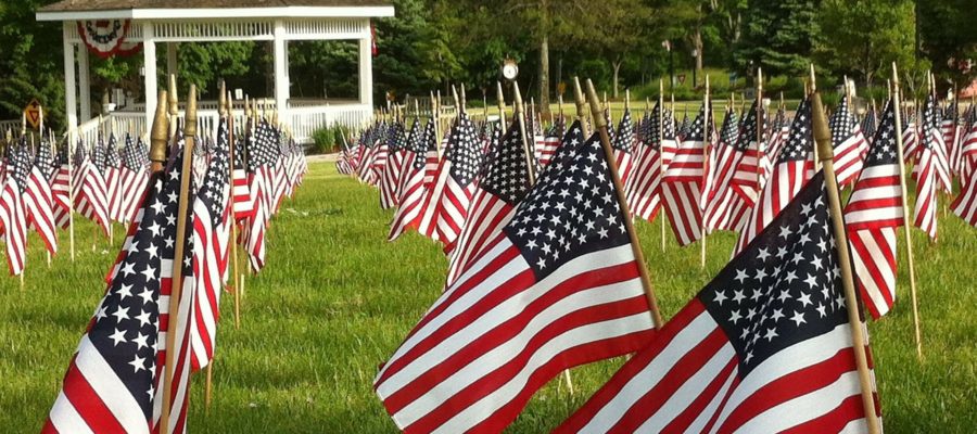 Small American flags arranged in rows on a grass lawn, near a white wooden gazebo with trees in the background