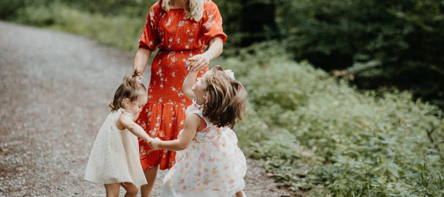 Mother in a red dres dances with two young girls in white dresses, on a gravel path among green foliage.